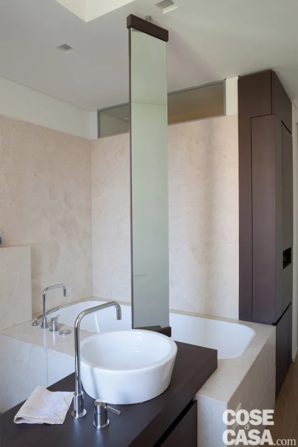 La vasca interpreta la vocazione wellness e relax della stanza. In questo caso, ciò che caratterizza il modello a incasso tradizionale è l'ampia struttura in muratura che lo sorregge, dimensionata in modo da formare un largo bordo laterale, utile come seduta oltre che come superficie d'appoggio. È interamente rivestita, come le pareti del locale, in lastre di pietra di Brera, un elegante marmo di particolare pregio, dal naturale colore bianco avorio, con diffusa granulometria grigio chiaro. Qui si innesta la base del lavabo, con un gioco di incastri dall'effetto scenografico al quale contribuisce lo specchio basculante fissato anche al top e al soffitto.