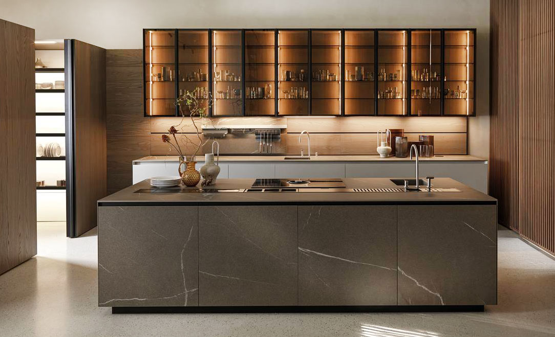 Nella cucina Telero di Euromobil si articola intorno all'isola con le basi in gres Laminam In-Side pietra piasentina taupé e attrezzata con il lavello e il piano induzione. Sulla parete di fondo sono appesi i pensili Teca con il telaio testa di moro e vetro fumé con una struttura in olmo Michelangelo come la boiserie attrezzata Horizon e le colonne Antis. Il pensile Teca misura L 45 x  H 120 cm, prezzo 856 euro + IVA. https://www.euromobil.com