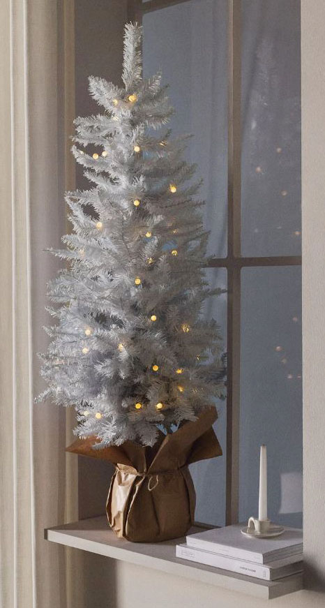 Può essere sistemato anche davanti ad una finestra l'abete James di Sklum che ha dimensioni contenute e una colorazione bianco candido. È completato da piccole luci gialle tra i rami e ha il basamento racchiuso con carta da pacchi e chiuso con un cordino. Misura Ø 60 x H 123 cm. Prezzo 32,95 euro. https://www.sklum.com