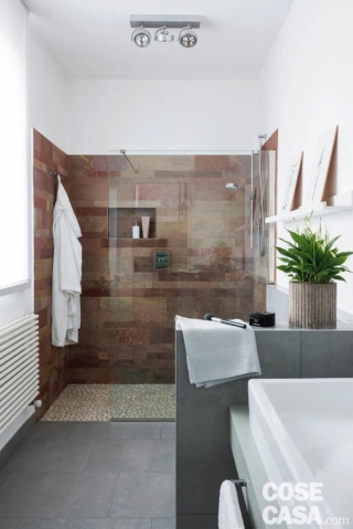 Questo bagno di grandi dimensioni presenta un soffitto molto alto e risulta ben illuminato dalla presenza di un'ampia finestra sul lato lungo. La luminosità, accentuata dal bianco delle pareti, è smorzata dalla presenza di elementi scuri, quali il colore grigio del gres porcellanato del pavimento e del rivestimento del muretto e dal colore scuro delle lastre in pietra che rivestono la doccia walk-in. Il locale non ha bisogno di una particolare illuminazione aggiuntiva e come lampadario è stata scelta una struttura costituita da tre faretti orientabili montati su una plafoniera in metallo cromato, che risulta quindi lineare ed essenziale.
Progetto d'interni Anna Marras, Milano http://www.annamarras.it - Foto Cristina Galliena Bohman - Styling Laura Mauceri