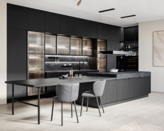 Si apre completamente sulla zona giorno la cucina Kalì di Arredo3 che è dominata dalla grande isola dotata di tavolo da pranzo con l’elegante struttura a ponte. Le ante delle basi sono in Laminato Rovere Fumé, i pensili e le colonne sono in Pet riciclato Stromboli. La zona lavaggio a parete è incorniciata da colonne e pensili con ante Teca in vetro millerighe e ripiani in vetro completati dalle luci a Led Back. Il Laminam Nero Greco dei piani di lavoro è ripreso dallo schienale Duetto attrezzabile con gli accessori magnetici Origami e dotato di illuminazione. Prezzo su richiesta. https://www.arredo3.com