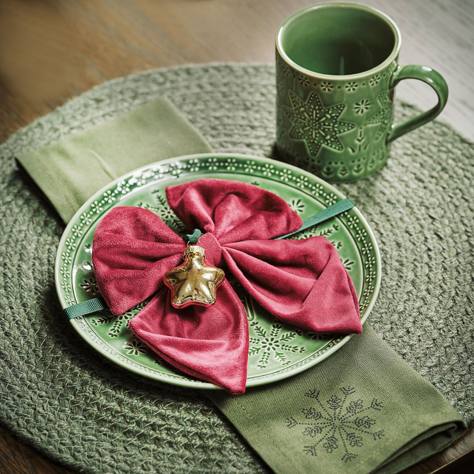 Una raffinata sfumatura verde oliva per il posto tavola per le feste di Jysk che è composto dal  piatto Malakit in porcellana lucida con un motivo naif di stelle e fiocchi di neve, dalla tazza coordinata e dalla tovaglietta rotonda Flinsten in iuta verde con un aspetto rustico. Il piatto misura Ø 20 cm, prezzo 5,50 euro. La tazza misura Ø 8 x H 9 cm, prezzo 4 euro. La tovaglietta misura Ø 38 cm, prezzo 5 euro. Il grande fiocco in velluto rosso con la stellina dorata funge da segnaposto. https://jysk.it