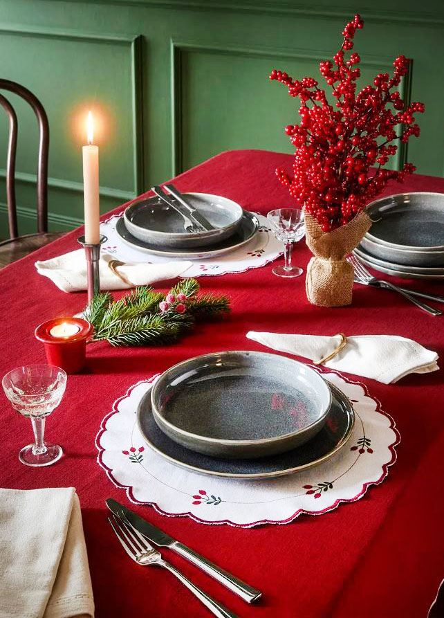 Le tovagliette americane rotonde (Codice articolo: 91835495) di Bonprix, realizzate in puro cotone biologico bianco con delicati ricami floreali rossi e verdi stile retrò, sono posizionate come segnaposto sopra una tovaglia rossa tinta unita.  Misurano Ø 38 cm, prezzo 14,99 euro (per il set da 2 pezzi). Sono abbinate a  stoviglie grigie e a bicchieri in vetro trasparente. Come centrotavola è stato usato un alberello con rami e bacche rosse.  https://www.bonprix.it