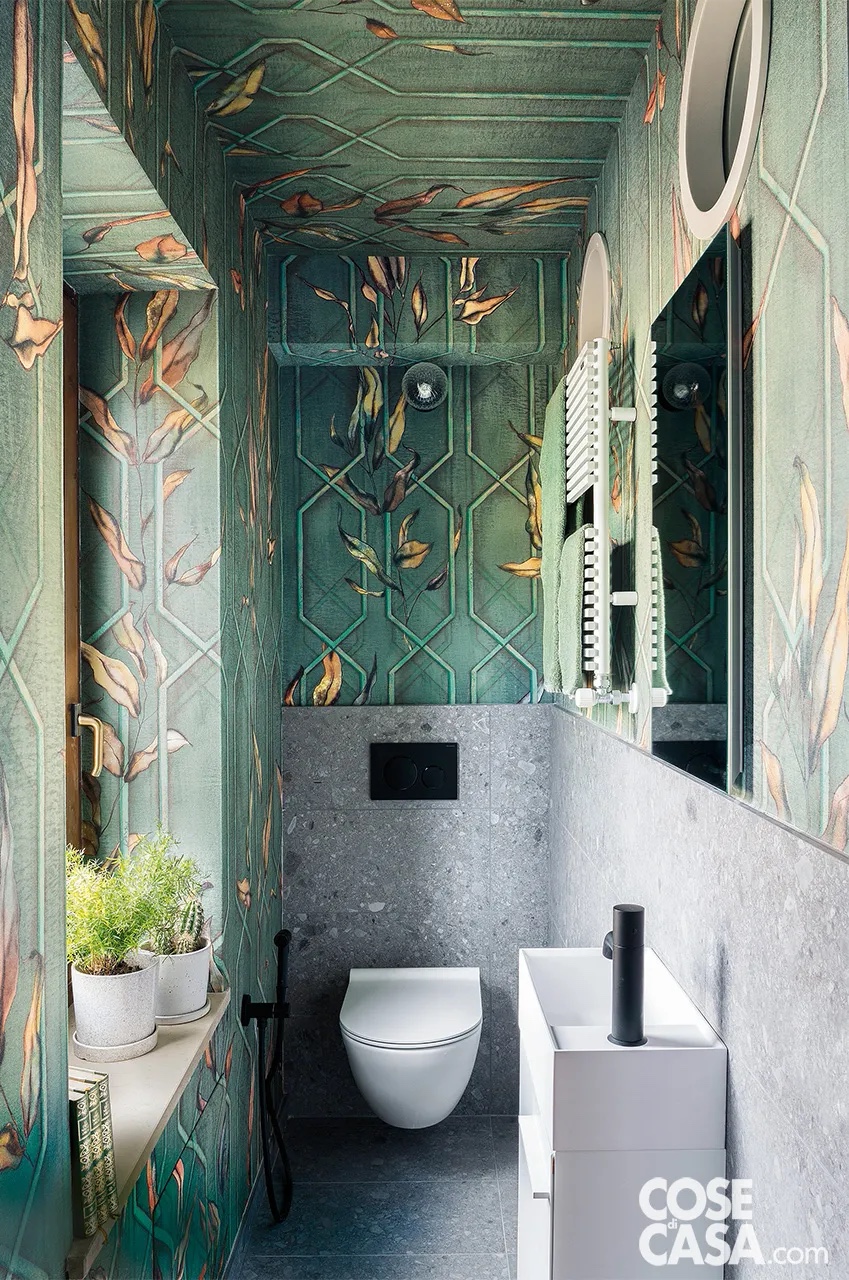 In questo bagno molto piccolo la tappezzeria a fondo verde con disegni ispirati alla natura ricopre interamente la parete con la finestra e il soffitto, mentre parte da metà muro nelle altre due pareti che ospitano lavabo e sanitario sospeso. Qui da terra a metà altezza il muro è protetto da un rivestimento in gres effetto pietra, uguale al pavimento, che sembra quasi una boiserie: il risultato è un bagno particolare ed elegante.
Progetto: arch. Maurizio Toffol, Via R. Koch, Fiera di Primiero (Tn) - Progetto d’Interior: Christian Benini - Foto: Alessandra Ianniello - Styling: Officina Interior