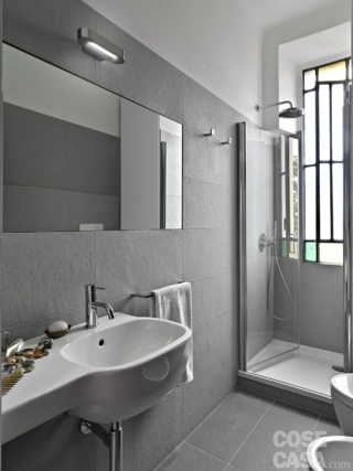 In questo bagno lungo e stretto, di soli 3,5 mq, sono state scelte sia per le pareti sia per il pavimento delle grandi lastre di gres effetto pietra, di colore grigio e dalla superficie ruvida e opaca, posate in orizzontale. La piastrellatura arriva in tutto l'ambiente fino a un'altezza di 200 cm, sia lungo le pareti lunghe sia nella doccia in muratura, in cui si trova la finestra, in corrispondenza della quale il rivestimento è più basso, limitato al muretto sotto l'apertura.
La parte superiore del bagno, invece, è dipinta di bianco come il soffitto. Il risultato è di grande semplicità e uniformità.
Progetto: arch. Tommaso Giunchi – Milano https://www.tommasogiunchi.it - Foto: Adriano Pecchio