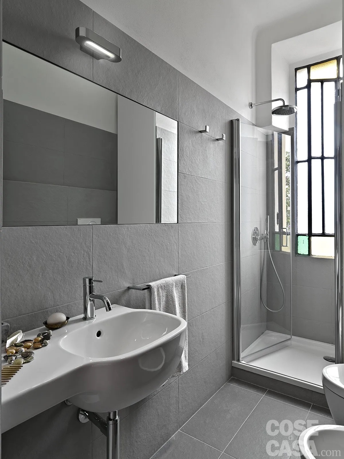 In questo bagno lungo e stretto, di soli 3,5 mq, sono state scelte sia per le pareti sia per il pavimento delle grandi lastre di gres effetto pietra, di colore grigio e dalla superficie ruvida e opaca, posate in orizzontale. La piastrellatura arriva in tutto l'ambiente fino a un'altezza di 200 cm, sia lungo le pareti lunghe sia nella doccia in muratura, in cui si trova la finestra, in corrispondenza della quale il rivestimento è più basso, limitato al muretto sotto l'apertura.
La parte superiore del bagno, invece, è dipinta di bianco come il soffitto. Il risultato è di grande semplicità e uniformità.
Progetto: arch. Tommaso Giunchi – Milano https://www.tommasogiunchi.it - Foto: Adriano Pecchio