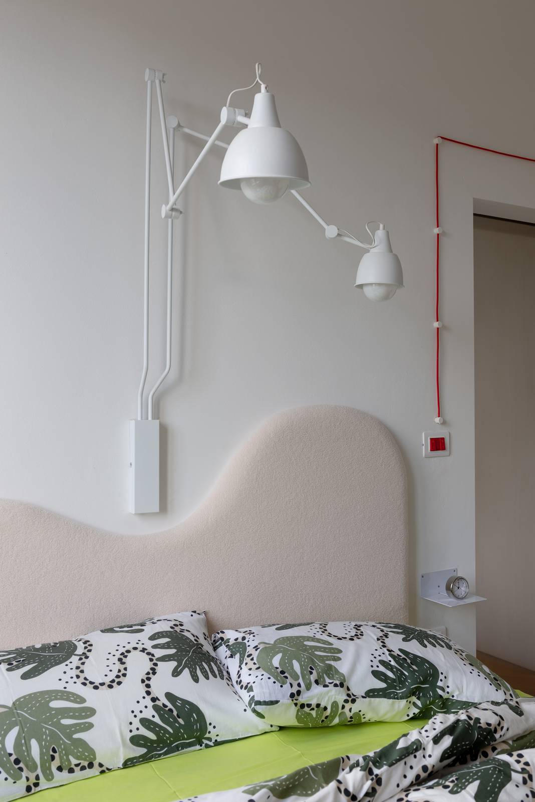 Testiera del letto imbottita in tessuto rosa cipria con profilo asimmetrico; sopra applique da parete a doppio braccio estensibile in metallo bianco con due lampade orientabili. A destra, cavo elettrico a vista rosso che scorre lungo il muro fino all'interruttore. Testiera del letto imbottita in tessuto rosa cipria con profilo asimmetrico; sopra applique da parete a doppio braccio estensibile in metallo bianco con due lampade orientabili. A destra, cavo elettrico a vista rosso che scorre lungo il muro fino all'interruttore.