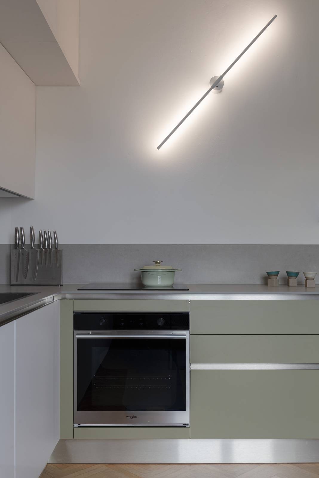 Dettaglio dell'angolo cottura con piano a induzione, piano di lavoro in ceramica e basi in verde salvia. Sulla parete una lampada a stelo lineare LED orientabile in metallo bianco, posizionata in diagonale. Forno da incasso in acciaio. Dettaglio dell'angolo cottura con piano a induzione, piano di lavoro in ceramica e basi in verde salvia. Sulla parete una lampada a stelo lineare LED orientabile in metallo bianco, posizionata in diagonale. Forno da incasso in acciaio.