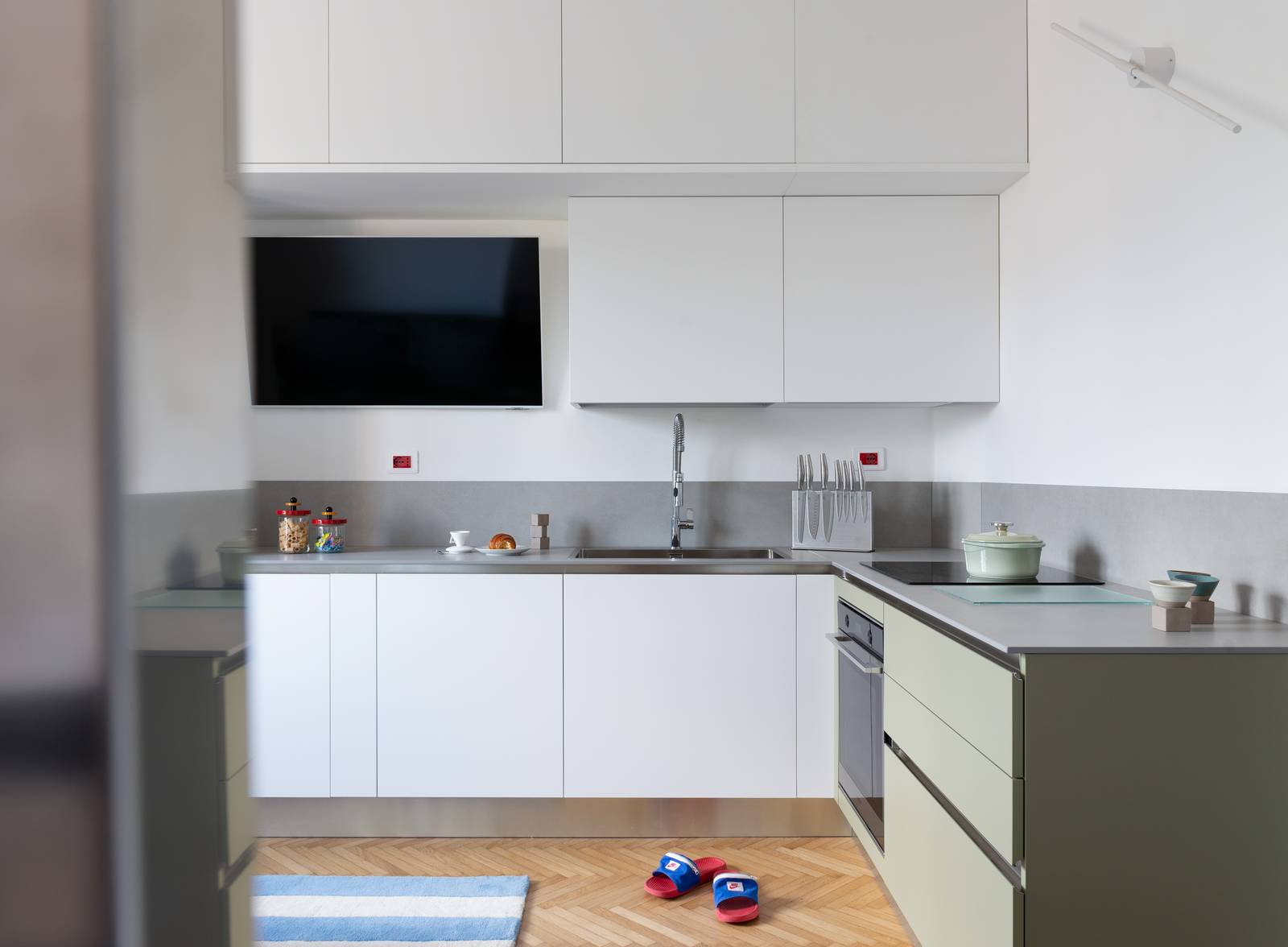 Vista d'insieme della cucina a L con basi e pensili bianchi, piano di lavoro in ceramica e alzatina abbinata. Angolo cottura con basi in verde salvia e forno da incasso. Televisore integrato nella parete. Sul pavimento in parquet a spina di pesce un tappetino a righe blu e bianche. Vista d'insieme della cucina a L con basi e pensili bianchi, piano di lavoro in ceramica e alzatina abbinata. Angolo cottura con basi in verde salvia e forno da incasso. Televisore integrato nella parete. Sul pavimento in parquet a spina di pesce un tappetino a righe blu e bianche.