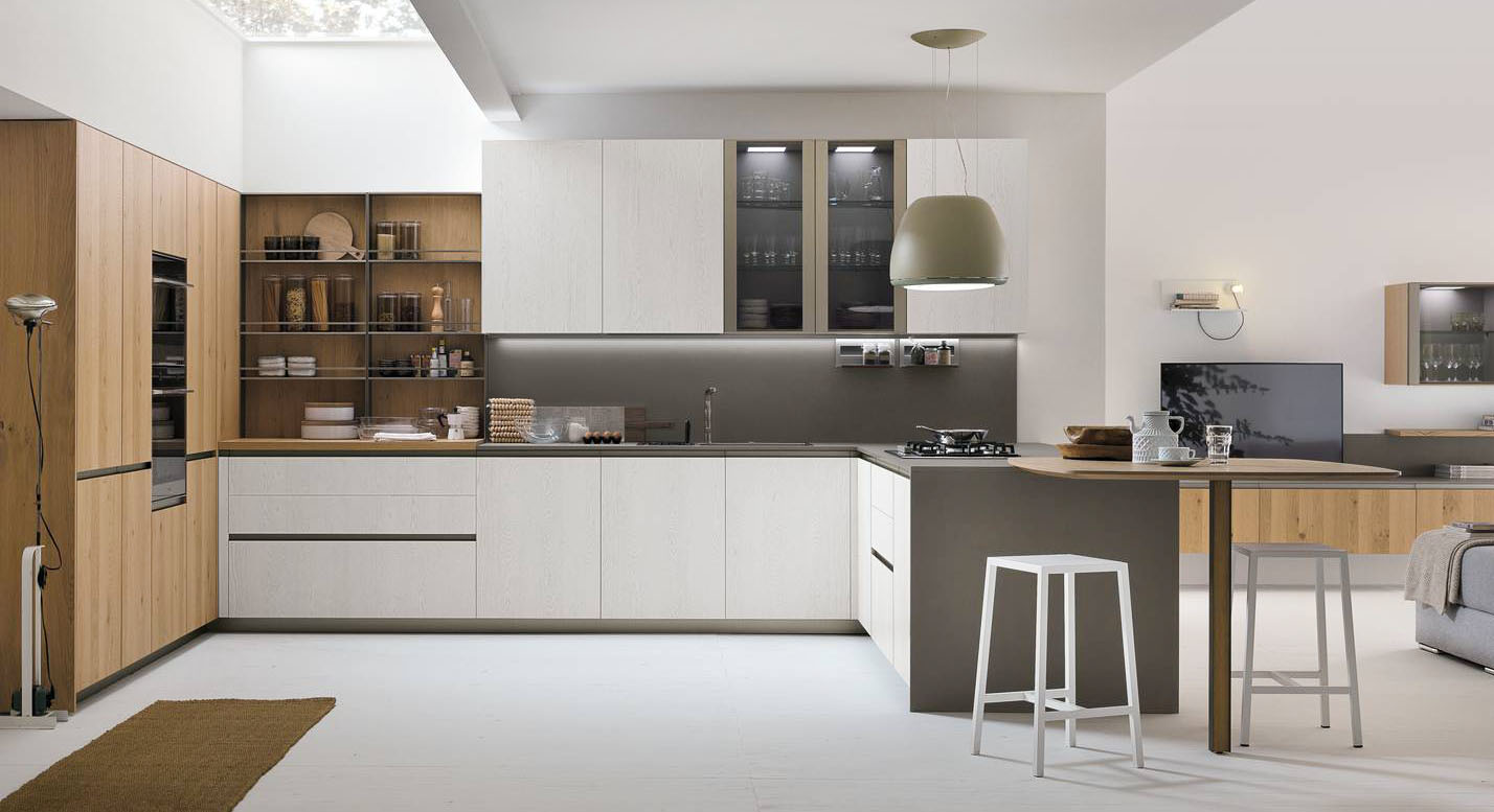 Si ispira allo stile nordico la composizione ad angolo Natural di Stosa che accosta le ante in Rovere Bianco a quelle in Rovere Miele. La penisola ha il top in pietra Neolith grigia, come lo schienale, e integra il piano snack, dalla forma squadrata, che permette di consumare pasti veloci e aumenta la superficie di lavoro a disposizione. I pensili con le ante in vetro permettono di esporre bicchieri e stoviglie come in una vetrina. Prezzo su richiesta. https://www.stosacucine.com