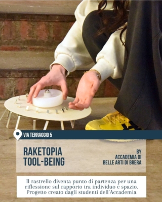 Fuorisalone 2026 Milano, 5VIE Design District: installazione “Raketopia Tool-Being” dell’Accademia di Belle Arti di Brera. Una persona sistema un oggetto circolare bianco simile a un piccolo tavolo-scultura, ispirato al rastrello, seduta sui gradini di uno spazio espositivo.