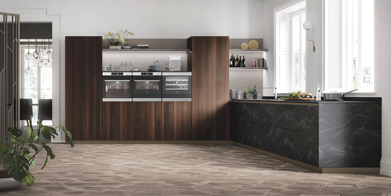 Si sviluppa lungo l'angolo la cucina AL 32 di Arrex che è caratterizzata da volumi compatti e dall'assenza di pensili. È composta da basi e colonne nella finitura Eucalipto Affumicato e da una serie di basi laterali con pannello HPL nella finitura effetto marmo scuro con venature a vista; le mensole in Fenix Grigio Londra con luce led integrata, che si inseriscono nello schienale con la stessa finitura, completano la composizione. Un modulo base da 60 cm, prezzo a partire da 746 euro + Iva. https://www.arrex.it