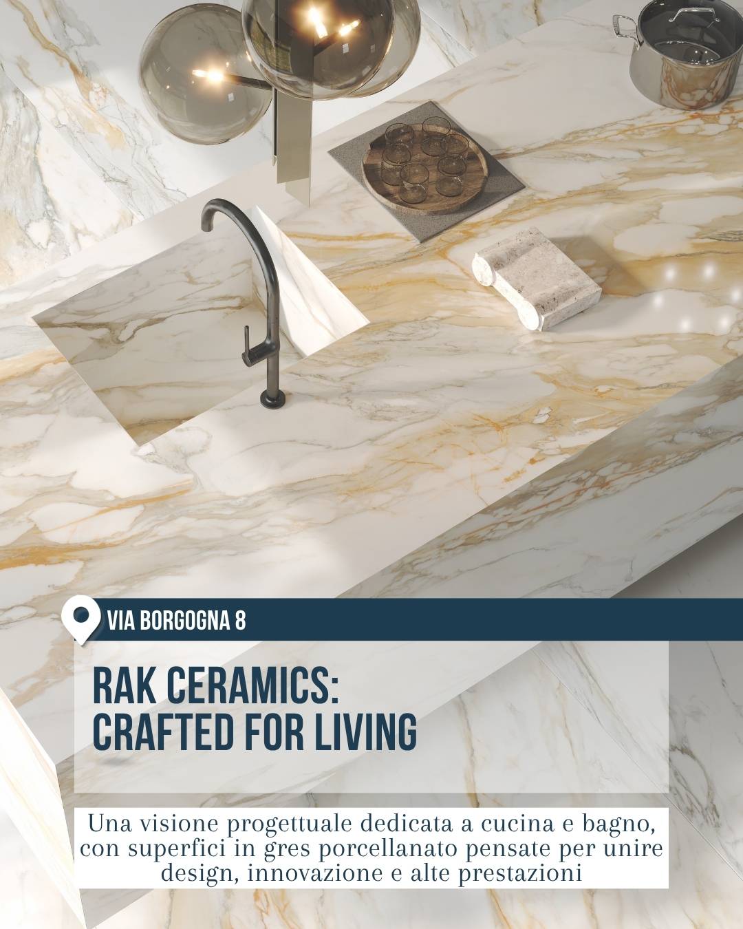 RAK Ceramics, showroom del Fuorisalone 2026 in via Borgogna 8 a Milano. Cucina contemporanea con top, penisola e lavello integrato in gres porcellanato effetto marmo bianco con venature beige e dorate, abbinati a rubinetto nero e lampade sospese.