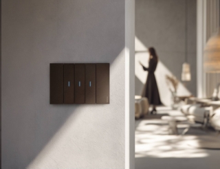 A modern living interior with plaster walls and soft materials. In the foreground, a flush-to-wall light switch plate in matte metal. A person in the background holds a smartphone, interacting with the environment. The light is natural and directional, entering from the side, creating strong contrast and sharp architectural shadows across the space. Subtle elements of the living area a sofa edge, a lamp, or a table complete the composition. Cinematic, intelligent, minimal. --ar 2:1 --v 7 Job ID: bc4c2457-7e98-4ca2-a235-6fa514c013dc