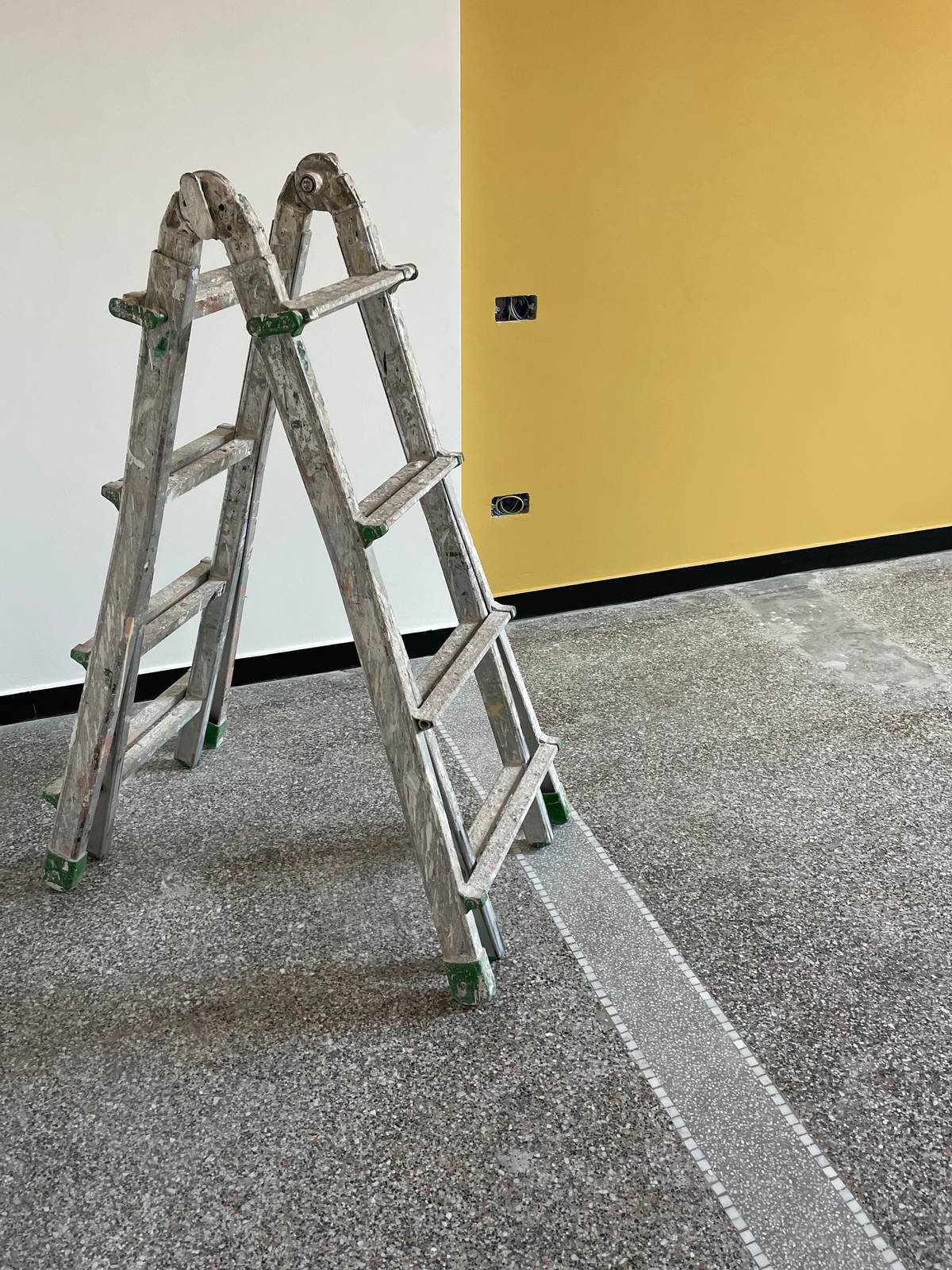 Nel restauro di questo pavimento originale in graniglia, la demolizione di un tramezzo ha lasciato una fascia centrale scoperta. Per conservare continuità con la superficie esistente, il rappezzo è stato trasformato in un elemento decorativo: una striscia in graniglia grigia delimitata da pietrine bianche che rende visibile l’intervento senza cercare di mimetizzarlo. Progetto architetto Clara Bona