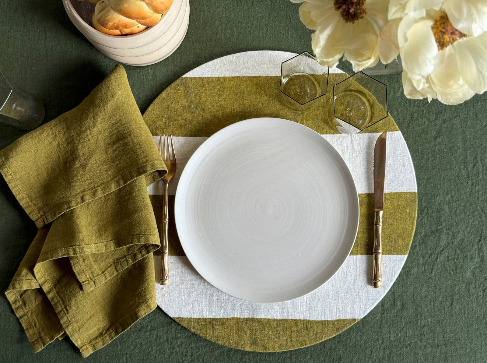 Il copritovaglietta rotondo di Once Milano è realizzato in lino decorato da un motivo a righe cabana verde matcha con effetto dipinto a mano. È rimovibile e necessita di un supporto interno in sughero; inoltre, è disponibile in due formati. Misura Ø 32 cm. Prezzo 56 euro per il set da 2 pezzi.  https://oncemilano.com