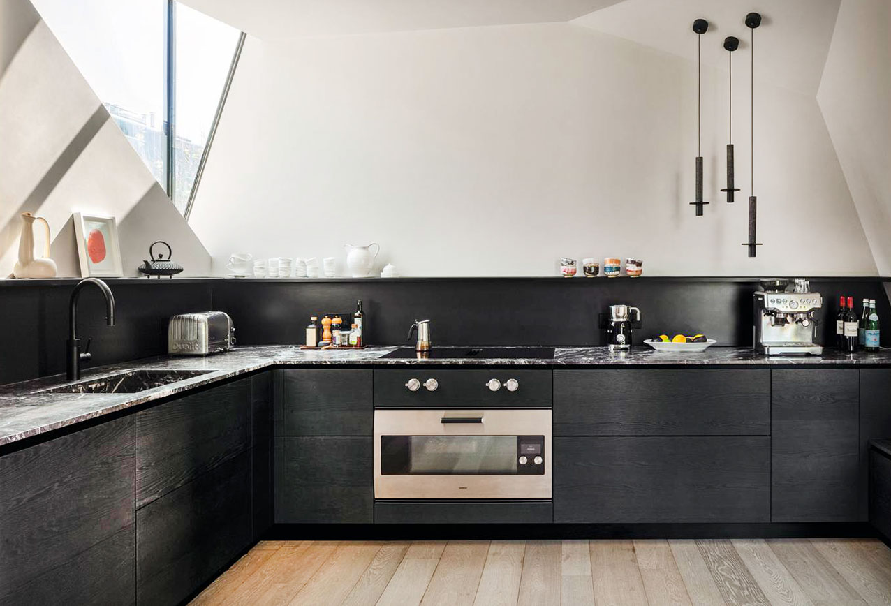 Priva di pensili la cucina ad angolo di Valcucine che è composta da una serie di basi con le ante in legno Olmo Tattile Nero, caldo e materico, e dal piano in pregiato marmo Grigio Carnico levigato con il lavello integrato nella stessa finitura. Il sistema di apertura con gola traccia un segno grafico che separa e al tempo stesso valorizza i materiali. https://www.valcucine.com