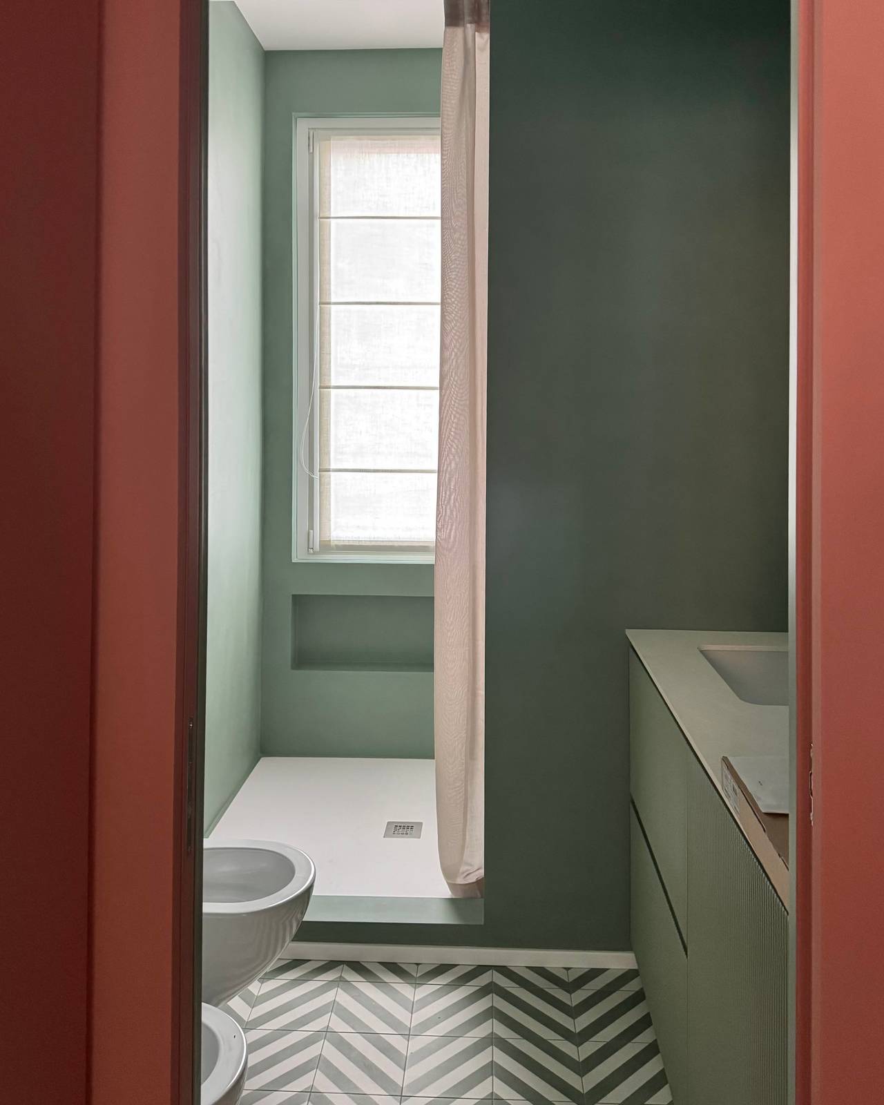 In questo bagno caratterizzato dall’uso del colore verde la doccia, con il piatto tagliato a misura realizzato in una miscela di quarzi, rimane posizionata sotto la finestra e separata dal resto del bagno grazie a un muretto laterale. Sotto la finestra è stata creata una nicchia utile per riporre shampoo e prodotti. Prima del muro della doccia c’è il lavabo a incasso con armadietto che contiene anche la lavatrice, nascondendola completamente.