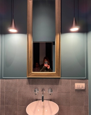 Ecco la dimostrazione di una verticalità voluta e enfatizzata: il lavandino è una catinella di dimensioni contenute, della stessa larghezza dello specchio con cornice dorata che sale in altezza fino quasi a toccare il soffitto. A lato scendono due lampade a sospensione, che calano dall’alto invece delle tradizionali applique a parete, per completare l’illuminazione in maniera più insolita.