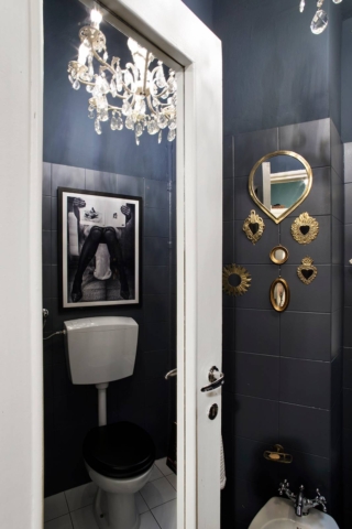 Un piccolissimo bagno dedicato esclusivamente alla zona dei sanitari, separati da lavandino e doccia. Le pareti sono state tutte dipinte di un intenso blu notte, quasi nero, sia quelle rivestite in piastrelle che quelle a intonaco. Per aggiungere una nota speciale sono stati aggiunti molto accessori scelti ad hoc: sulle pareti c’è una piccola collezione di specchi con cornici dorate o in ottone, a soffitto c’è un lampadario in cristallo, e sopra alla cassetta di scarico esterna del WC è appesa una foto a tema in Bianco e Nero. Tutte scelte di carattere e lievemente controcorrente.