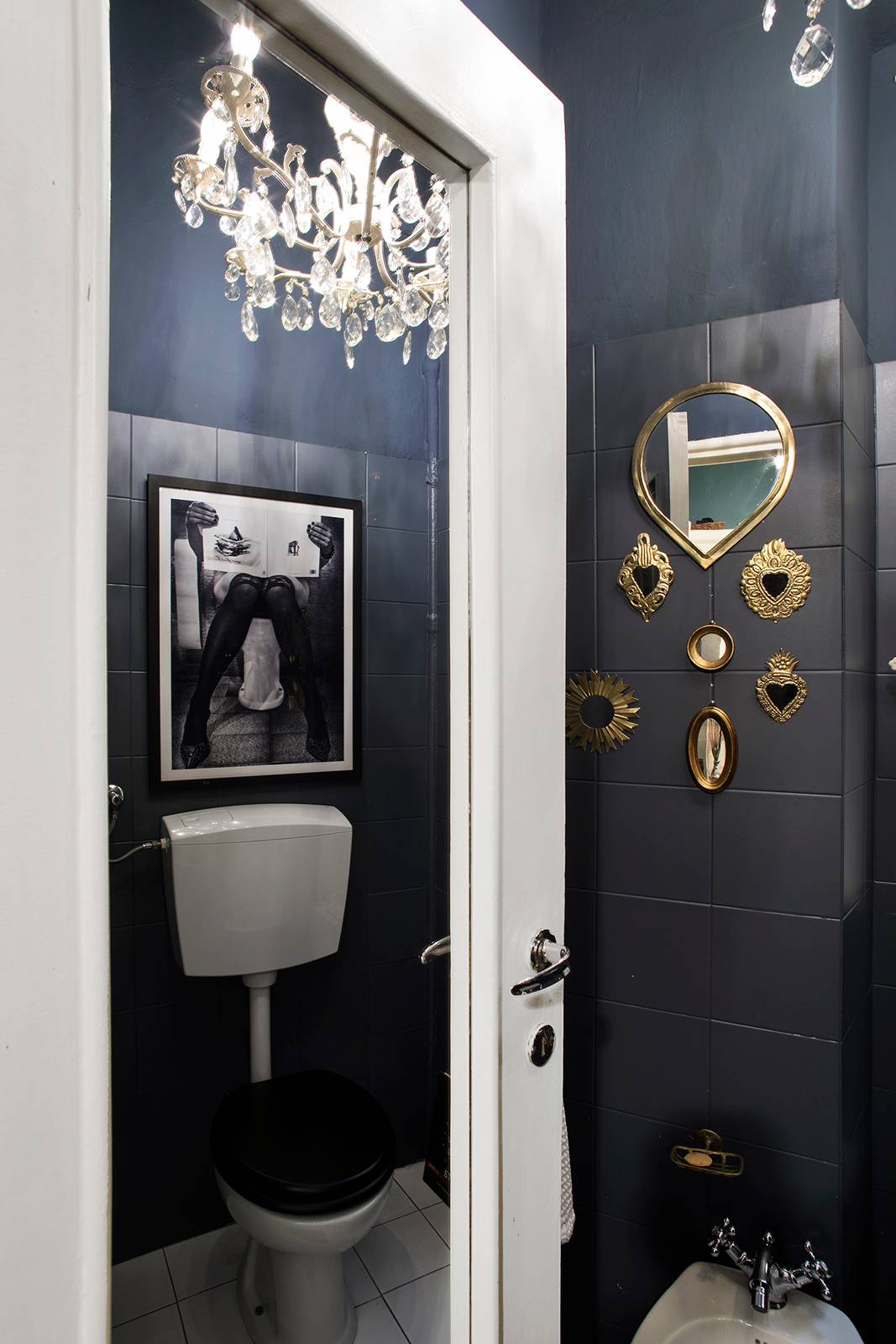 Un piccolissimo bagno dedicato esclusivamente alla zona dei sanitari, separati da lavandino e doccia. Le pareti sono state tutte dipinte di un intenso blu notte, quasi nero, sia quelle rivestite in piastrelle che quelle a intonaco. Per aggiungere una nota speciale sono stati aggiunti molto accessori scelti ad hoc: sulle pareti c’è una piccola collezione di specchi con cornici dorate o in ottone, a soffitto c’è un lampadario in cristallo, e sopra alla cassetta di scarico esterna del WC è appesa una foto a tema in Bianco e Nero. Tutte scelte di carattere e lievemente controcorrente.